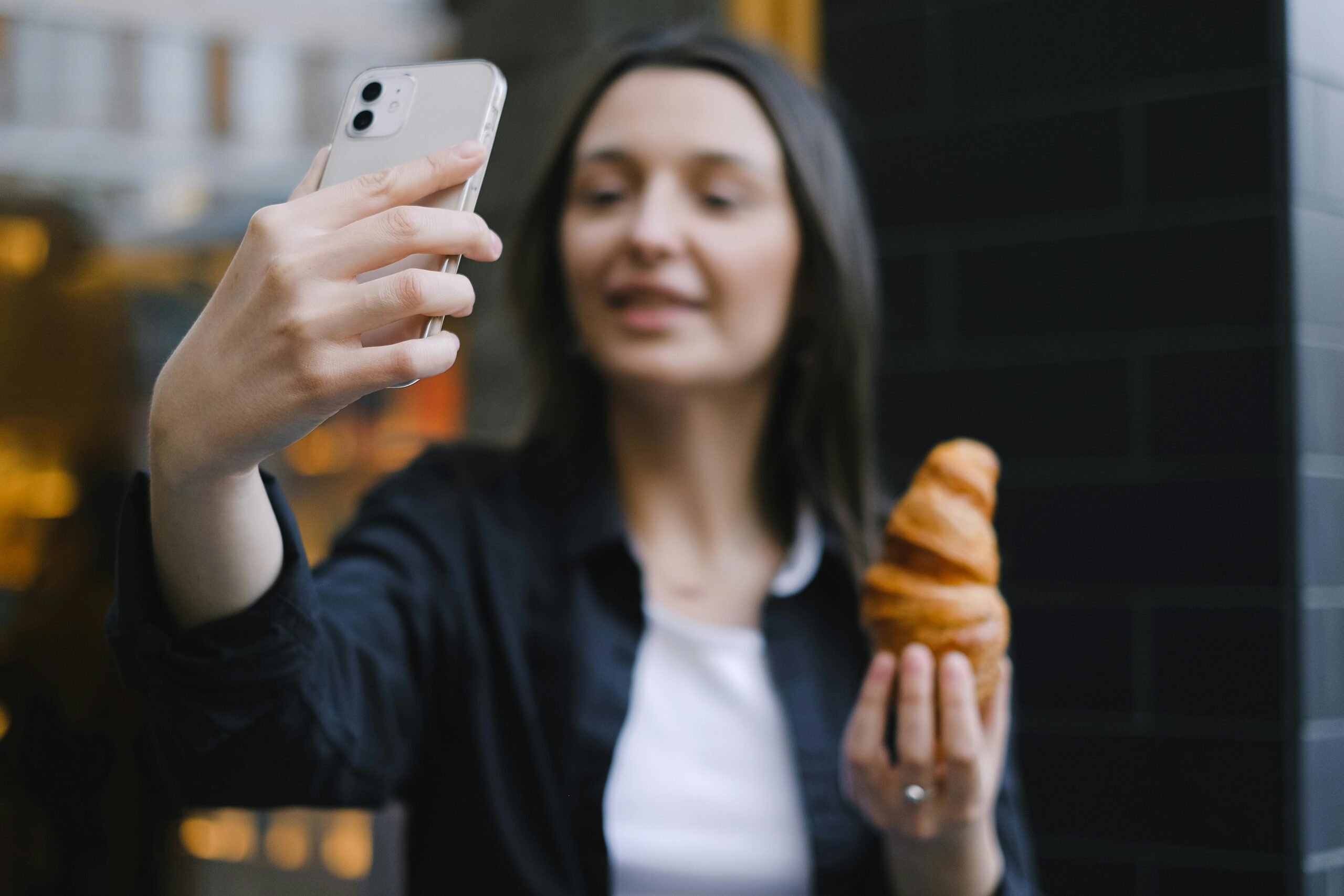 A woman takes a selfie while holding a croissant in an urban outdoor setting.
